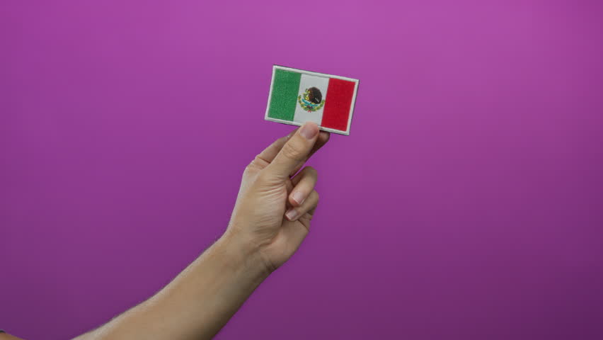 Man hand holding mexican flag embroidery against a vibrant pink wall, showcasing national pride and craft on an isolated colored background with emphasis on cultural textiles.