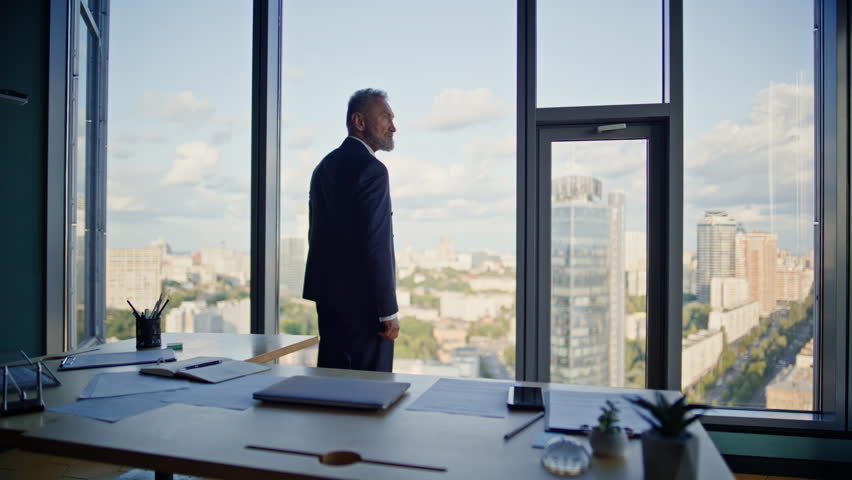 Business owner contemplating city view from modern office workplace. Senior confident businessman standing by panoramic window looking cityscape. Successful leader in elegant suit thinking of project.