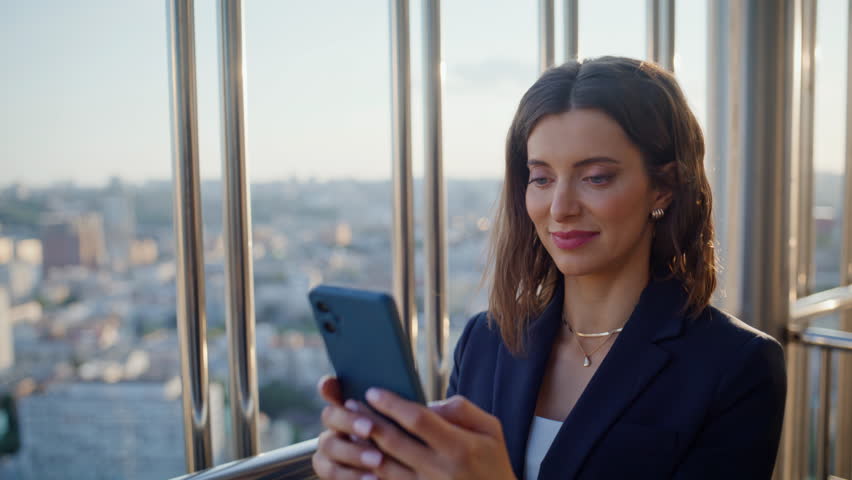 Cheerful businesswoman texting cellphone standing at balcony highrise building closeup. Smiling woman manager browsing phone banking app receiving salary. Happy employee reading message on terrace.