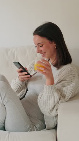 Vertical video, A young woman enjoys a relaxing morning at home, drinking orange juice and scrolling on her phone. She is happy and comfortable.