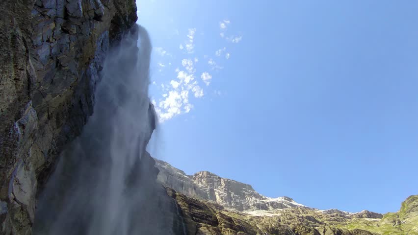 4K 60 fps horizontal shot of a majestic waterfall in the Pyrenees, cascading down rocky cliffs under a bright blue sky, showcasing natural beauty and mountain wilderness.