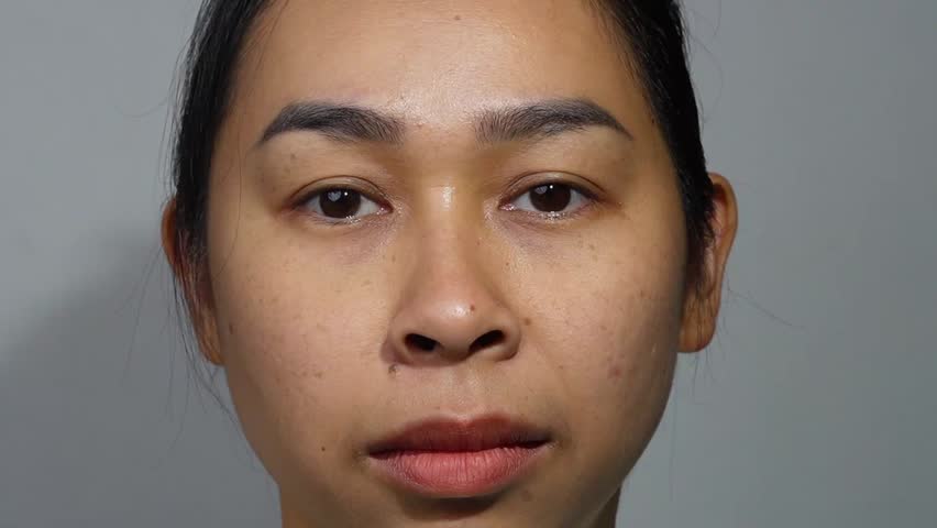 An Asian woman looking at camera with a thoughtful and slightly worried expression against a grey background.