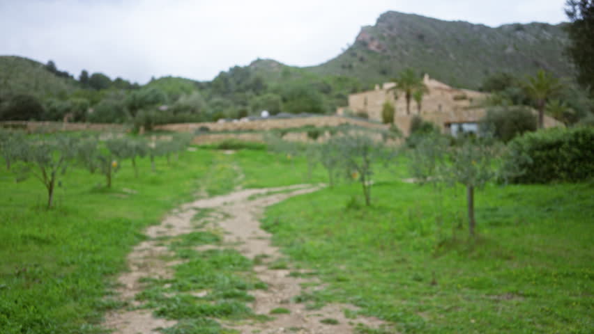 Blurred image depicting a defocused mediterranean landscape with green fields and a pathway leading to distant houses surrounded by trees and hills.