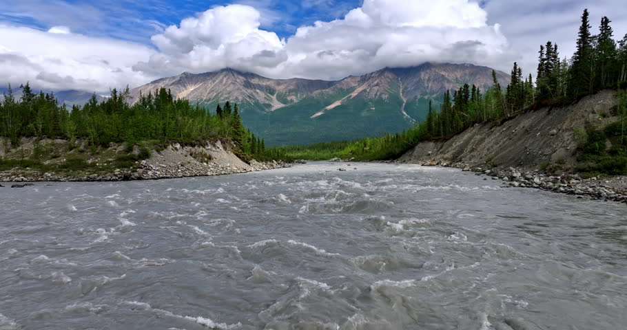 Turbulent mountain river with grey dirty water. Rocks with fluffy white cloudscape on top at backdrop. Alaska wilderness.