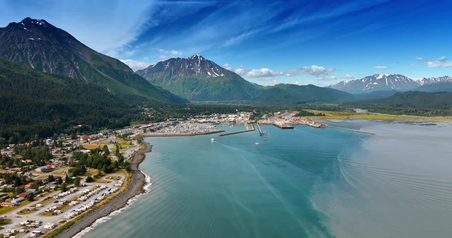 Flight over the blue waterscape of the lake near the town located on the beautiful waterfront. Evergreen forests and stunning mountains surround the urban area. Alaska, aerial view.