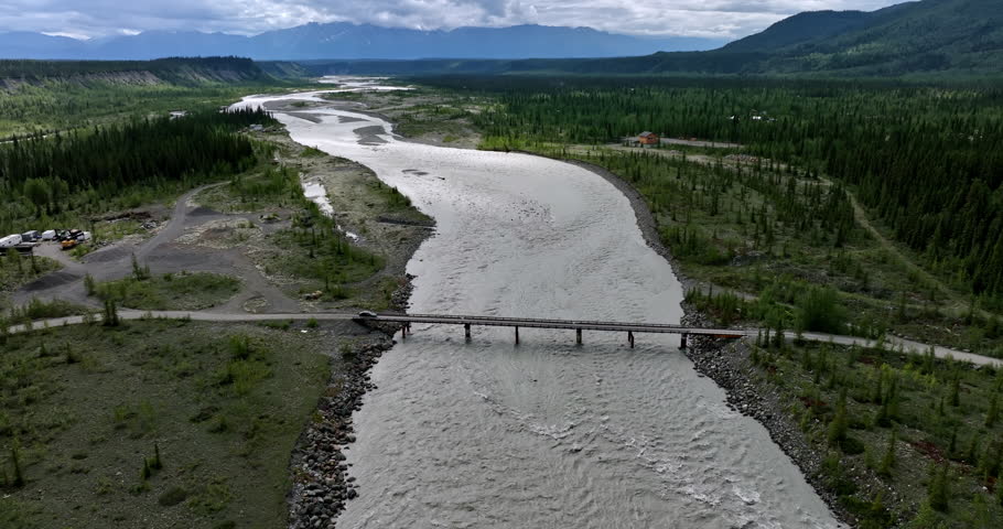 Approaching a bridge across the turbulent river crossed by the black car. Travel by the wild Alaska. Aerial view.