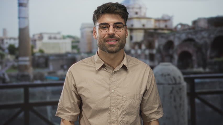 Hispanic man wearing eyeglasses and beige shirt making phone gesture in front of ancient roman building ruins; connection.