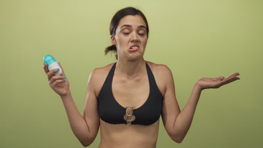 Woman holds roll on deodorant and shrugs with uncertain expression in olive green studio; personal hygiene dilemma.