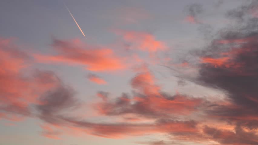 A plane flies through a colorful sky at sunset. The clouds are painted in shades of pink and orange, creating a serene atmosphere.