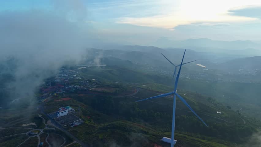 Aerial view of wind turbine rotating above scenic mountain landscape with fog on sunset.