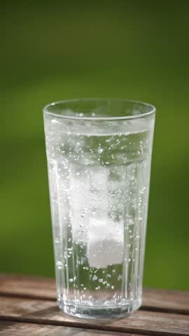 A human hand drops a soluble aspirin and vitamin C tablet into a glass of water, creating bubbles on a green summer garden background