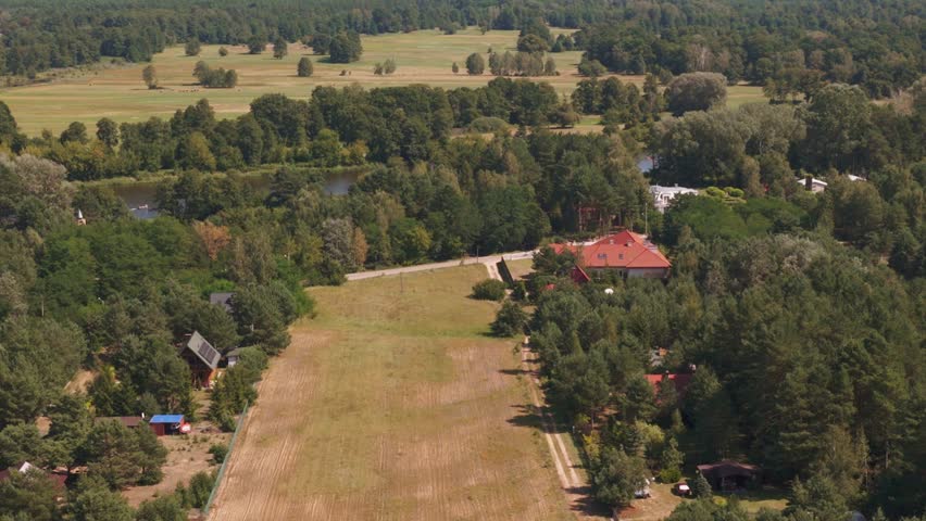 Drone captures green fields, dense forests, and a winding river in Nowy Lubiel, Poland. A red roofed building is visible amid the natural scenery.