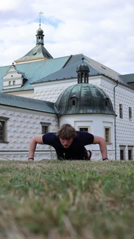 Vertical screen young man doing push ups on grass in front of historic European architecture combining outdoor fitness training with cultural city atmosphere discipline strength and healthy lifestyle