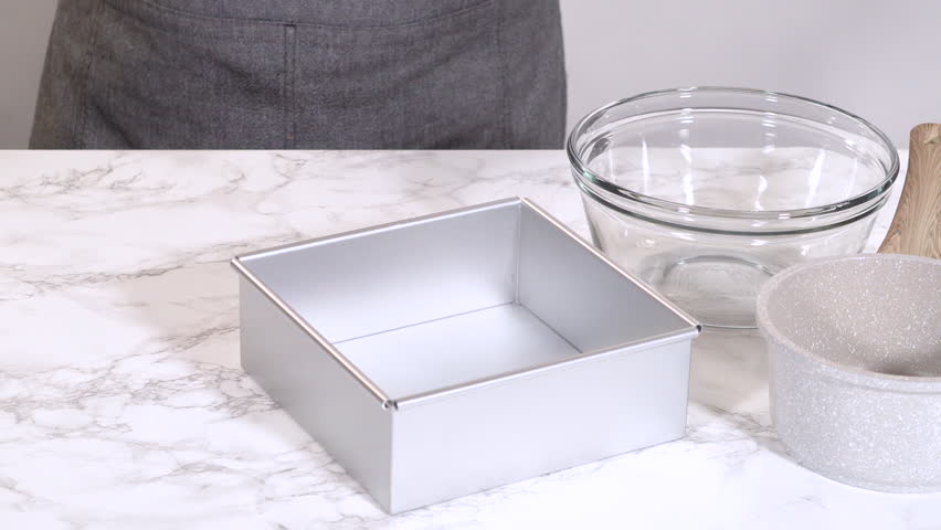 A metal baking pan, glass bowls, and a small saucepan are neatly arranged on a marble countertop. The setup is well-lit and organized, ideal for baking tutorials and kitchenware ads.