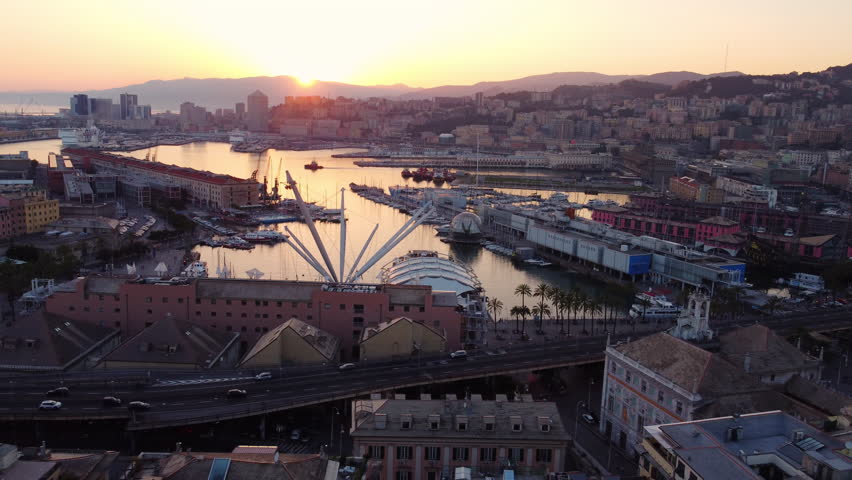 Drone flies backward and slightly upward over Genoa historic port at sunset, showing traffic, city, and mountains in the background.