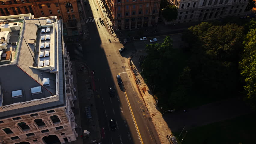 Drone top-down view of a Genoa street at sunset, with vehicles turning and half the road in sun, half in shadow.