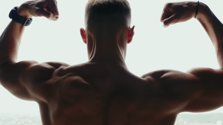 Rear view of a muscular man flexing his biceps, showcasing strength and fitness in a gym setting. The image emphasizes bodybuilding, masculinity, and dedication to physical health.