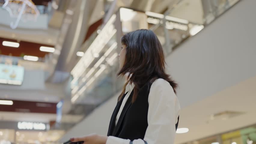 A young adult woman in casual business attire confidently walks in a bustling shopping center, suggesting a blend of lifestyle and modern ambiance. Confident Woman Walking Through a Shopping Mall