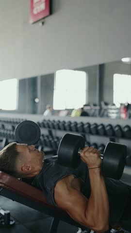 Side view of a young man performing a bench press with dumbbells in a gym, focused and determined. Young Man Doing Weightlifting Bench Press Exercise at the Gym. Vertical