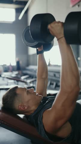 Side view of a young man performing a bench press with dumbbells in a gym, focused and determined. Young Man Doing Weightlifting Bench Press Exercise at the Gym. Vertical