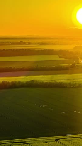 Bright orange sun setting over endless green crop fields, bathing the rural countryside landscape in a warm golden hour glow -vertical aerial time lapse