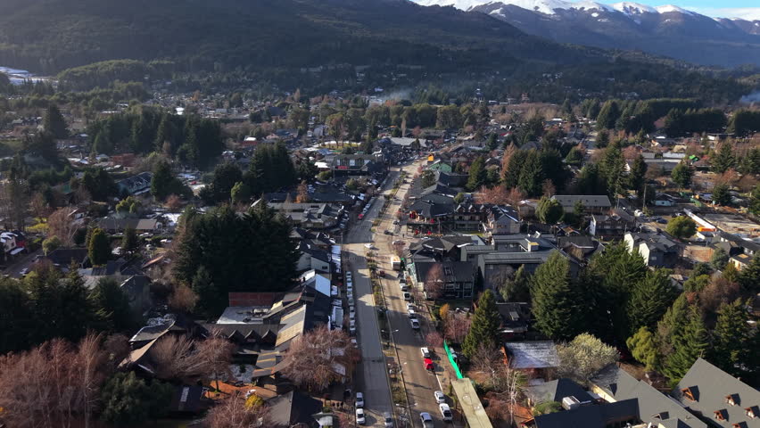 Aerial drone shot advancing over Villa La Angostura, Argentina, showing busy streets, traffic, and arriving tourists framed by forested hills and the Andes mountains.
