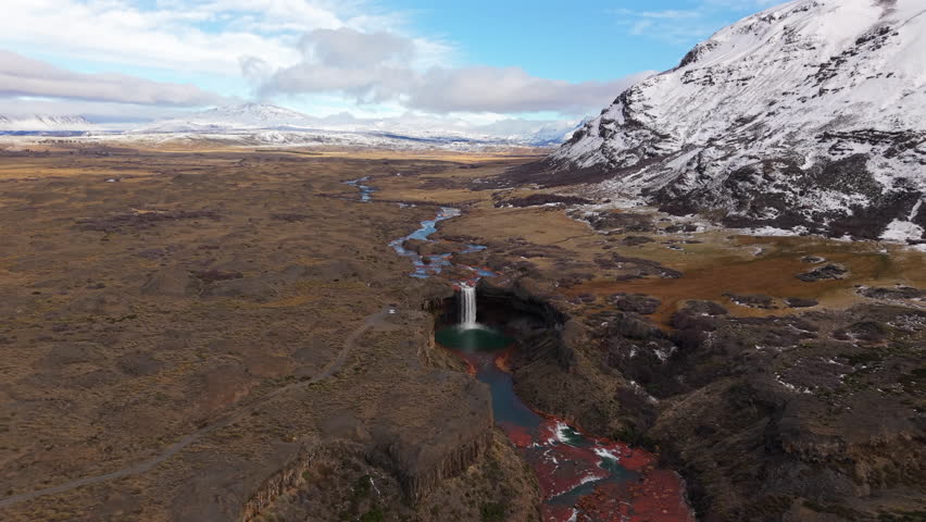 Drone pull away reveals Salto del Agrio in Caviahue, a 60m waterfall plunging into a green pool, framed by vivid basalt cliffs and ancient araucaria trees on the Patagonian Andes plateau.