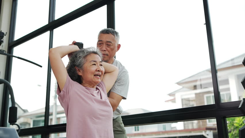 Asian Senior couple is working out together in a gym, focusing on stretching and strength exercises. Elderly Woman holds dumbbells while her husband encourages her, showcasing a healthy lifestyle.
