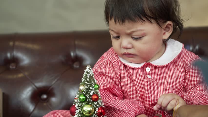 A baby girl in red dress playing with Christmas stuff on sofa, winter holiday season