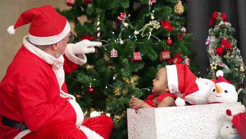 Santa Claus playing with a little girl in red dress sitting in a gift box. Represents Christmas magic, holiday joy, and childhood innocence