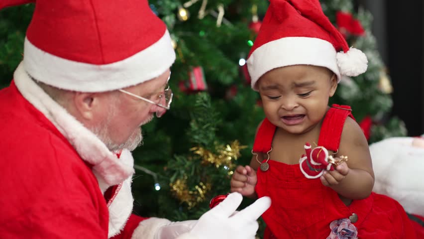 A senior man dress as Santa claus playing Christmas stuff with African baby in red dress at Christmas tree.
