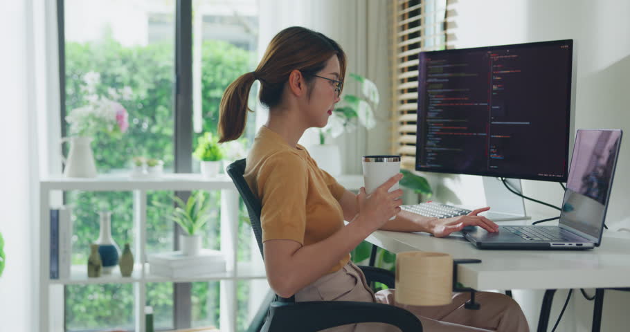 Young Asia female freelance eyeglasses drink coffee and type write prompt AI bot IT app program nomad, video game, terminal with coding, user interface, data center on desktop computer at home office.