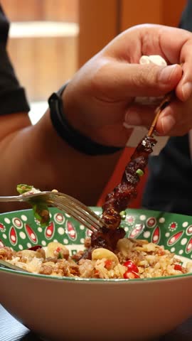Hand lowers grilled beef skewer onto rice meal in patterned bowl, with warm indoor lighting and close-up camera angle highlighting food texture