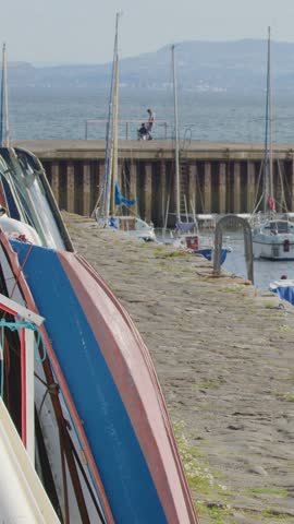 A person walks along a stone pier by the sea, with moored boats and a fishing village in bright daylight. Static camera, wide shot