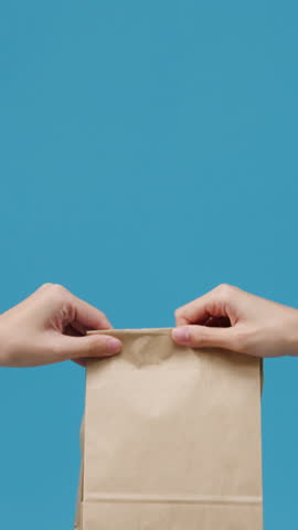 Vertical Screen : Young woman hand hold brown clear empty blank craft paper bag food for takeaway over blue background. Copy space for place text for advertisement. Advertising area, mock up content.