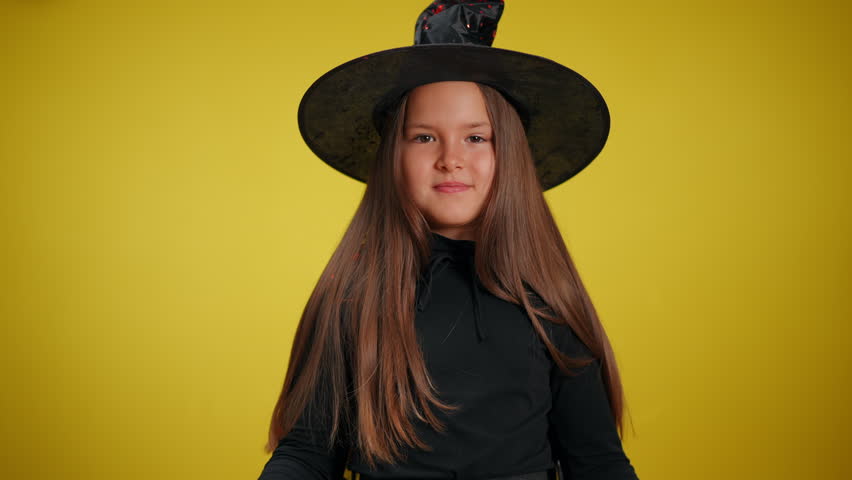 A young girl dressed up in a witch costume is playfully posing with her whimsical hat against a bright backdrop