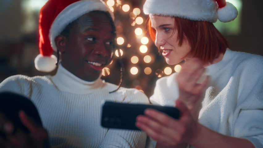 Wait for Christmas miracle, black man and Caucasian woman in Santa hats at home. Loving couple celebrating winter holidays together, using smartphone, watching funny video and pictures in internet