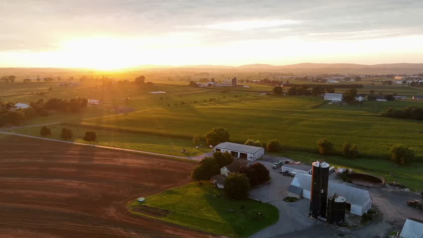Lens flare or golden sunlight in the evening. Aerial wide shot. America. Farmstead countryside with silos and barn: agricultural farm fields in summer. Golden sunset in USA.