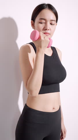 Vertical clip. Young sportswoman shows off her physical strength by lifting dumbbell on white background.