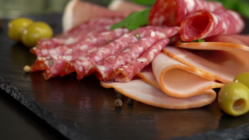 Close up of fresh slices cold cuts sausage ham bacon jamon rotate in circle on stone cutting board with olives and parsley. Meat. Pork beef. Food