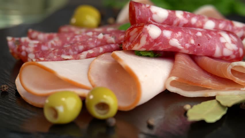 Close up of fresh slices cold cuts sausage ham bacon jamon rotate in circle on stone cutting board with olives and parsley. Meat. Pork beef. Food