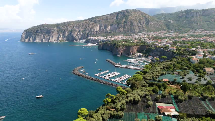 Aerial drone video of the Port of Piano di Sorrento, seen from the sea with the majestic tuff cliffs of the Sorrento coast overlooking the blue waters. A scenic view of the harbor, coastline, and Medi