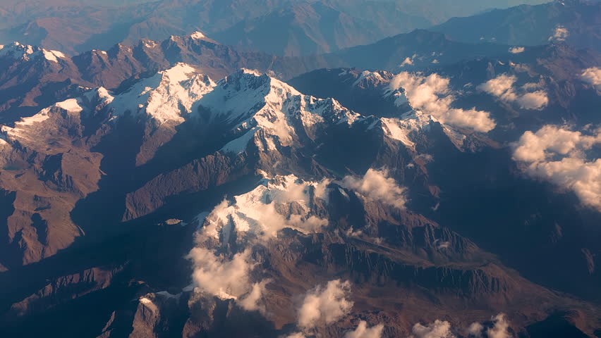 The majestic Andes mountains at sunset. The footage captures snow-capped peaks and stunning rock formations bathed in golden light, with clouds casting shadows below.