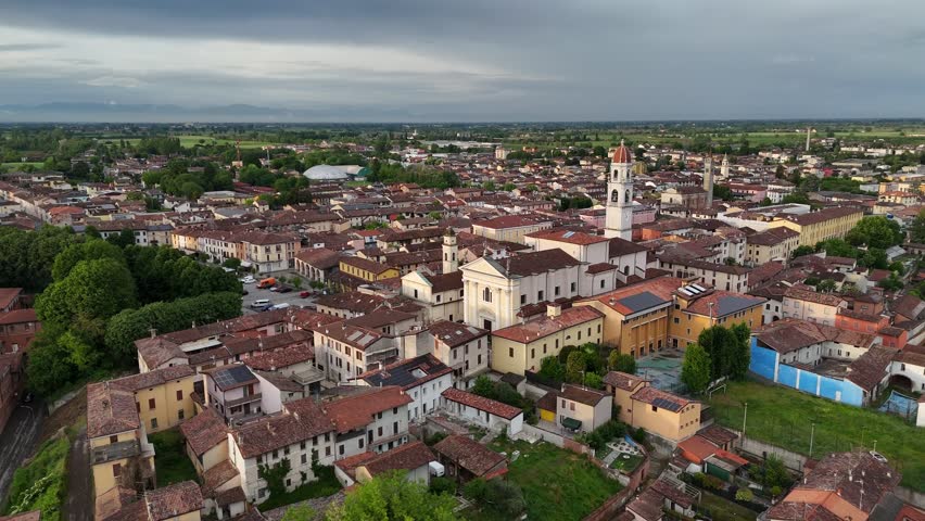 Drone orbit captures the historic Church of Santi Tommaso e Andrea Apostoli standing prominently in Pontevico, Brescia in Lombardy, near red-roofed houses and the flat northern Italian plain at dusk