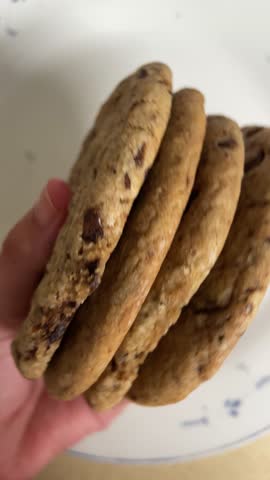 Stack of homemade chocolate chip cookies held by hand on a white plate, showing rustic texture and fresh baked dessert presentation.
