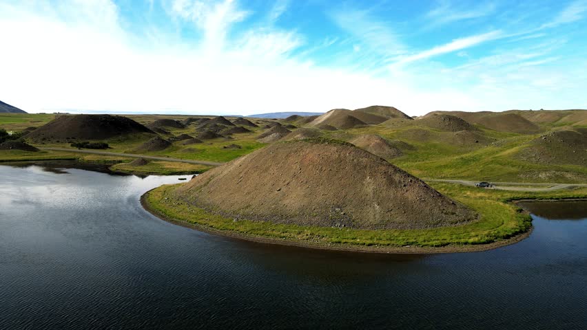 Aerial view of Vatsndalsvegur Road winding through a landscape of grassy mounds and a dark pond beneath a partly cloudy sky, Blondufos, Húnabyggo, Iceland.