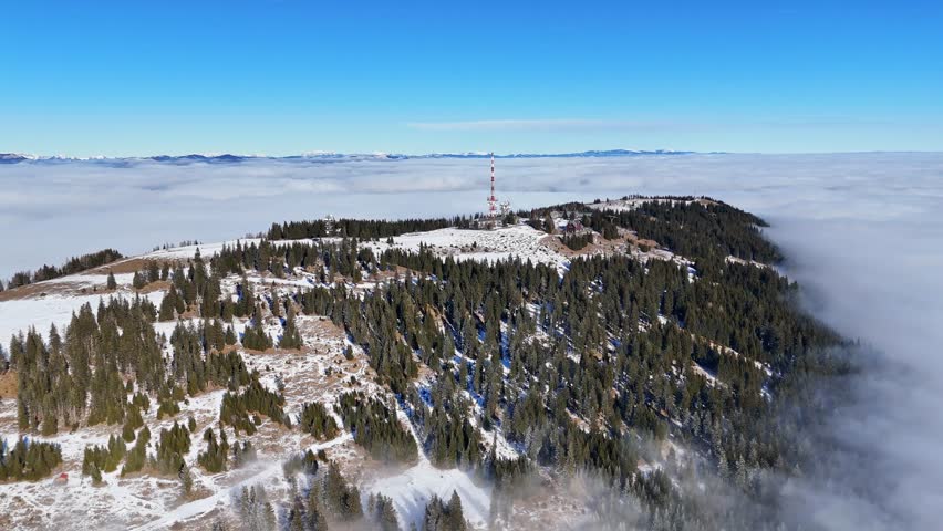 Aerial view of the snow-dusted Schockl mountain, crowned by a prominent tower, amidst a sea of clouds under a clear sky, Graz, Styria, Austria.