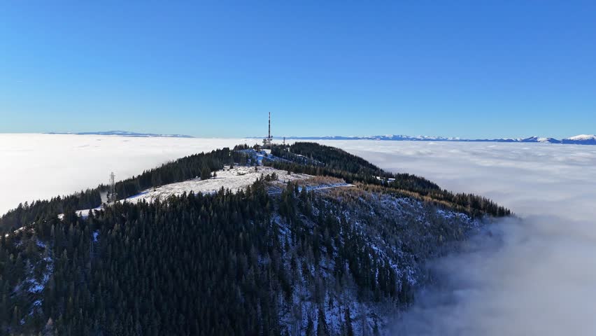 Aerial view of the Schockl mountain peak piercing through the clouds under a clear blue sky, creating a serene landscape, Graz, Styria, Austria.