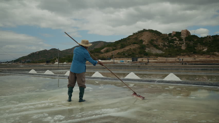 Old Asian man in straw hat harvests salt in evaporation pond in Vietnam. Elderly worker gathers mineral deposits with traditional rake on sea coast. Natural condiment manufacturing