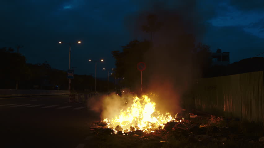 Large fire burns on place of car wreck on urban road edge at night. Bright flame and heavy smoke on broken automobile parts in evening urban environment. Safe driving importance